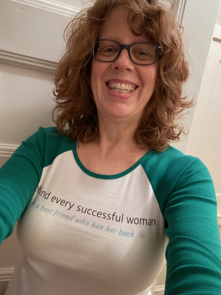Photo of Manya, smiling red-haired woman wearing a white and  green baseball style shirt with the words "Behind every successful woman is a best friend who has her back."